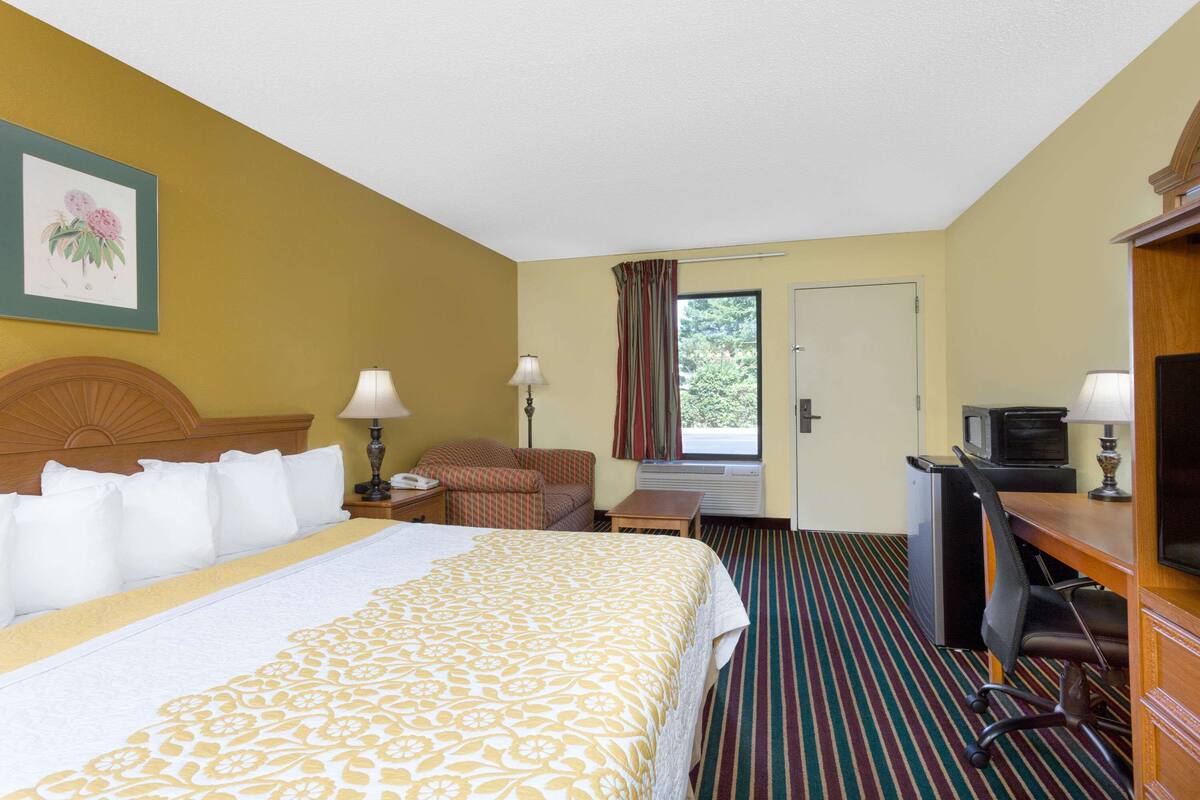 Quarto de hotel com uma cama, uma escrivaninha com televisão, uma cadeira, abajur e uma janela com cortinas.