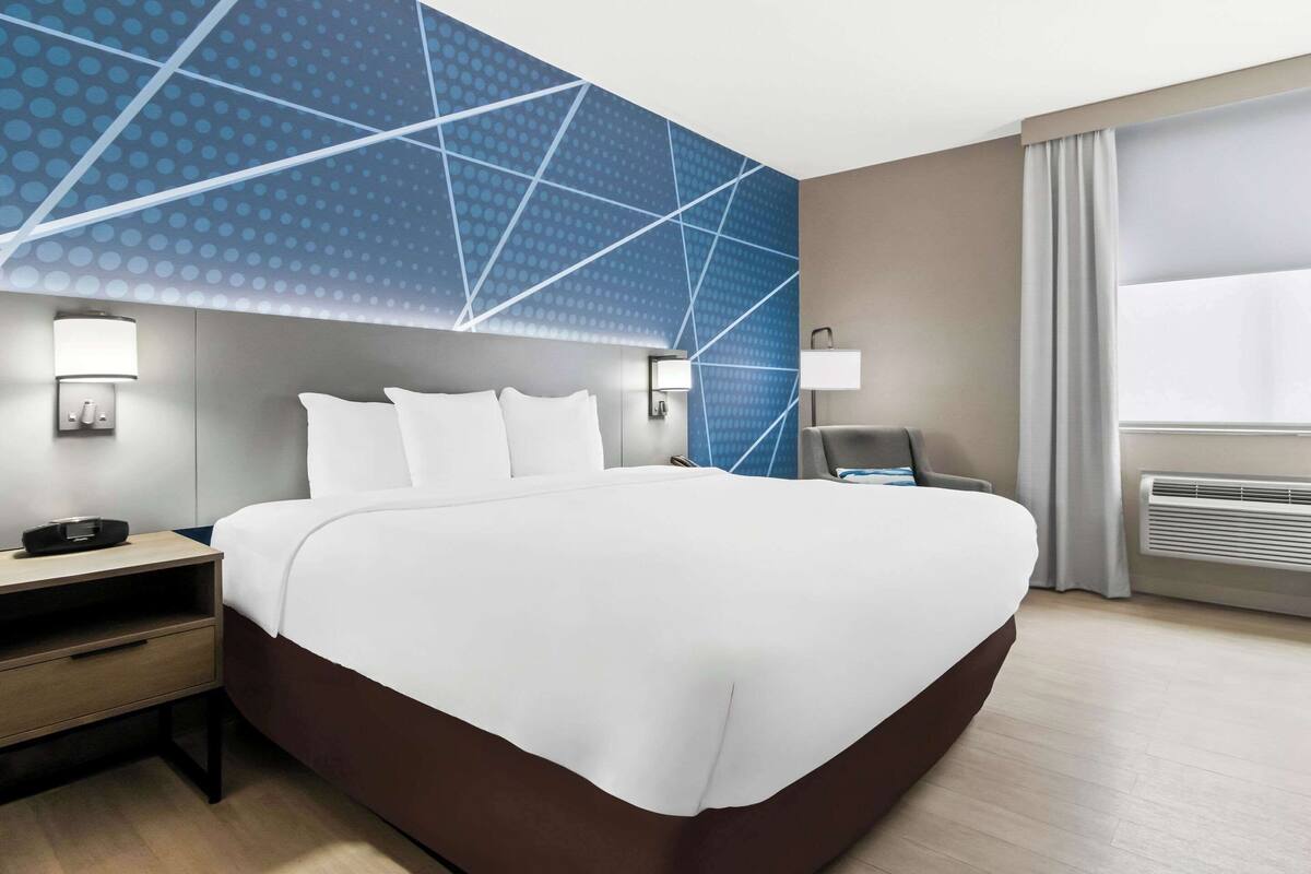 A modern hotel room with a large bed, a desk, a chair, and a window with curtains.