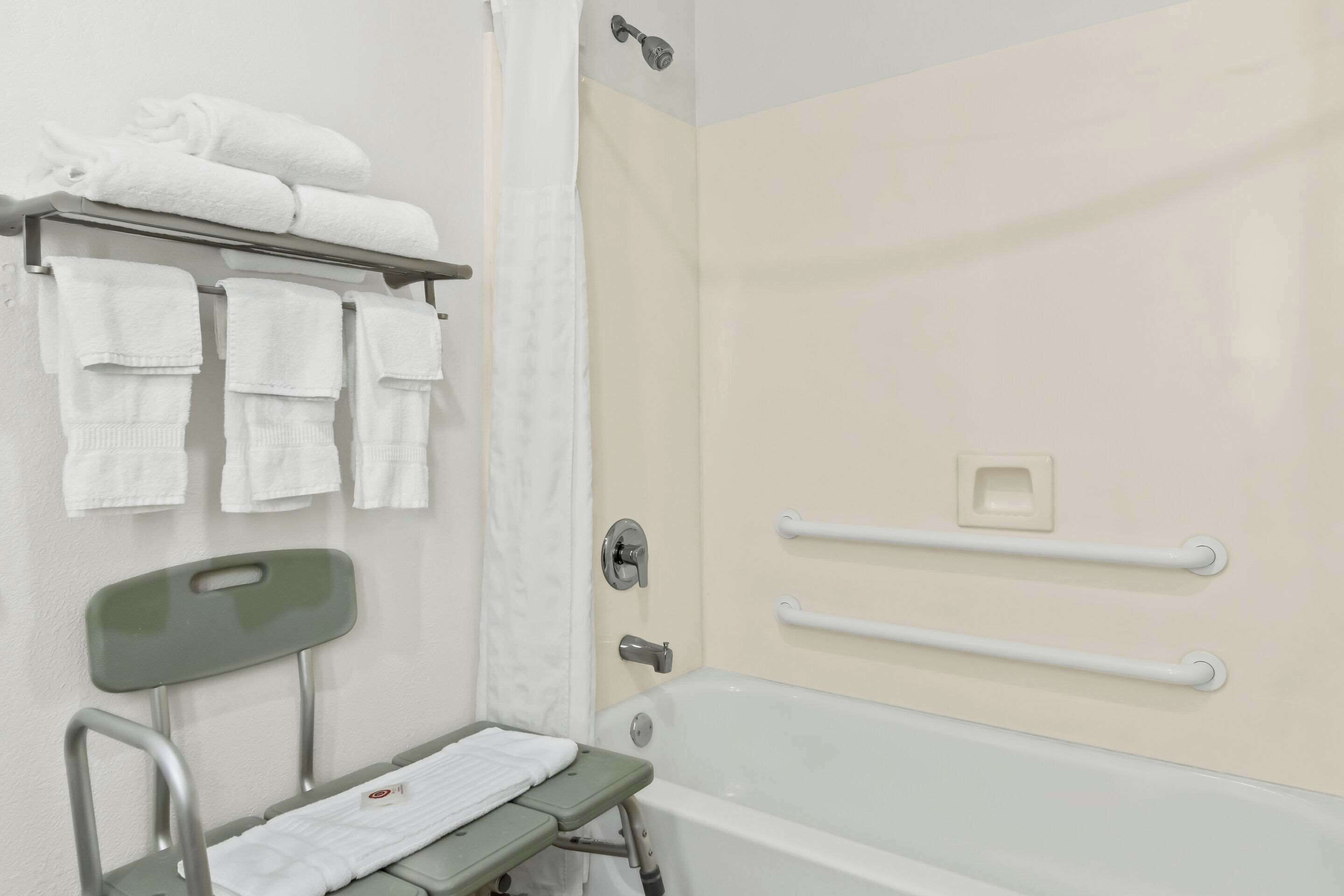 Un bagno con vasca, tenda da doccia, portasci con asciugamani, maniglia di sostegno e panca accessibile per sedia a rotelle.