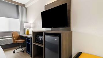 A modern hotel room with a flat-screen TV mounted on the wall, a desk with a chair, a microwave, and a refrigerator.