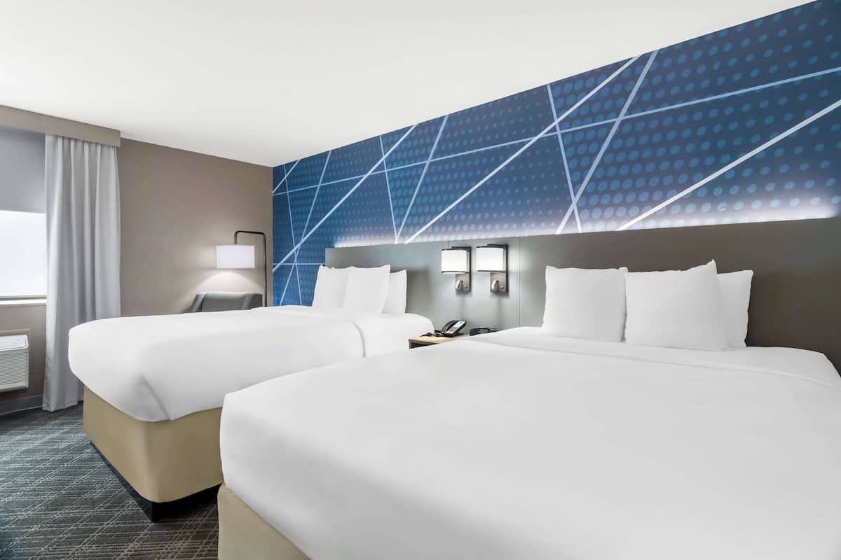 A hotel room with two beds, a large window with curtains, a wall-mounted lamp, and a geometric-patterned wall.