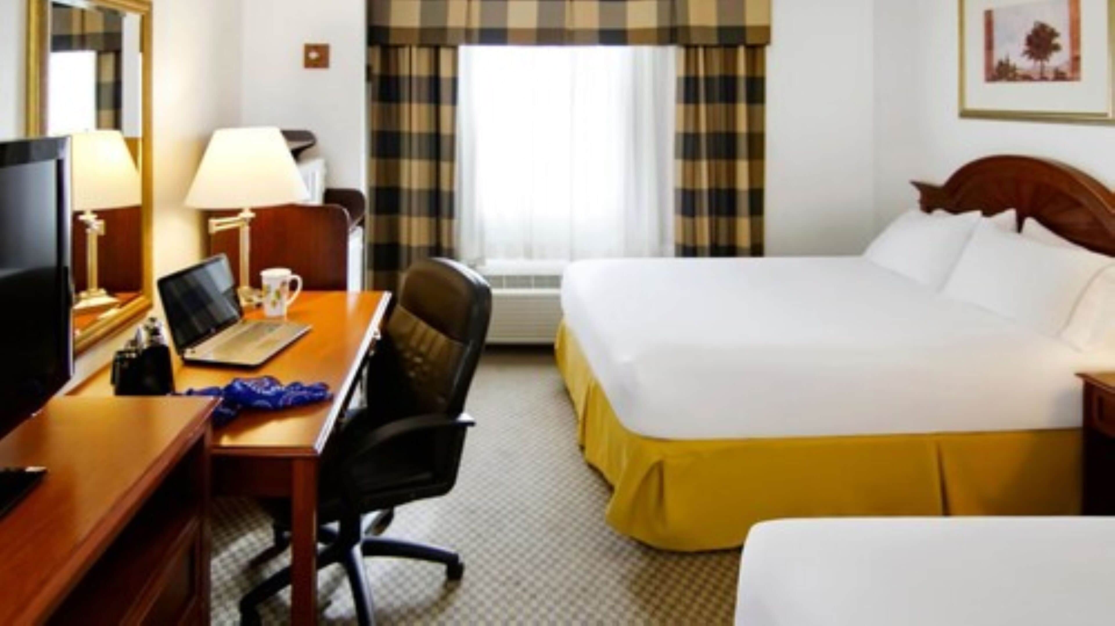 A hotel room with a bed, a desk with a laptop, a chair, a TV, and a window with curtains.