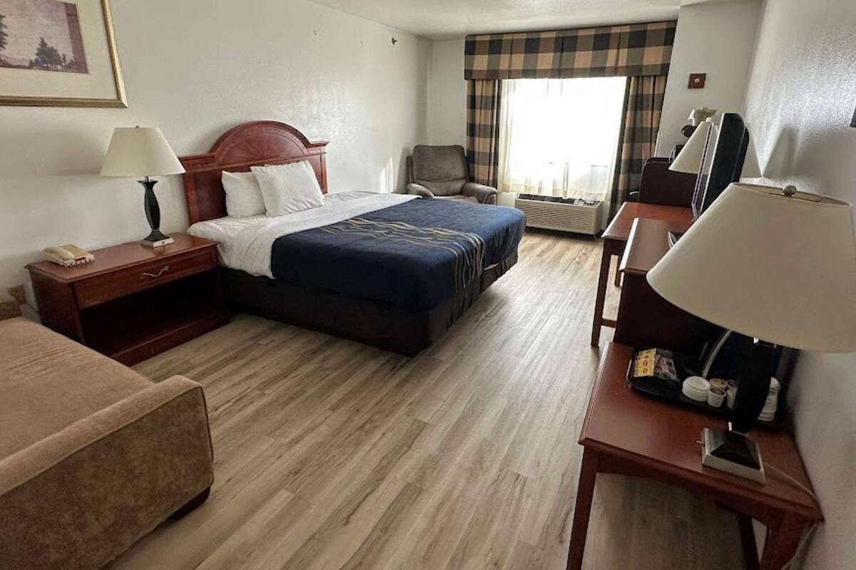 Ett hotellrum med en säng, sängbord, en stol, ett skrivbord och ett fönster med gardiner.