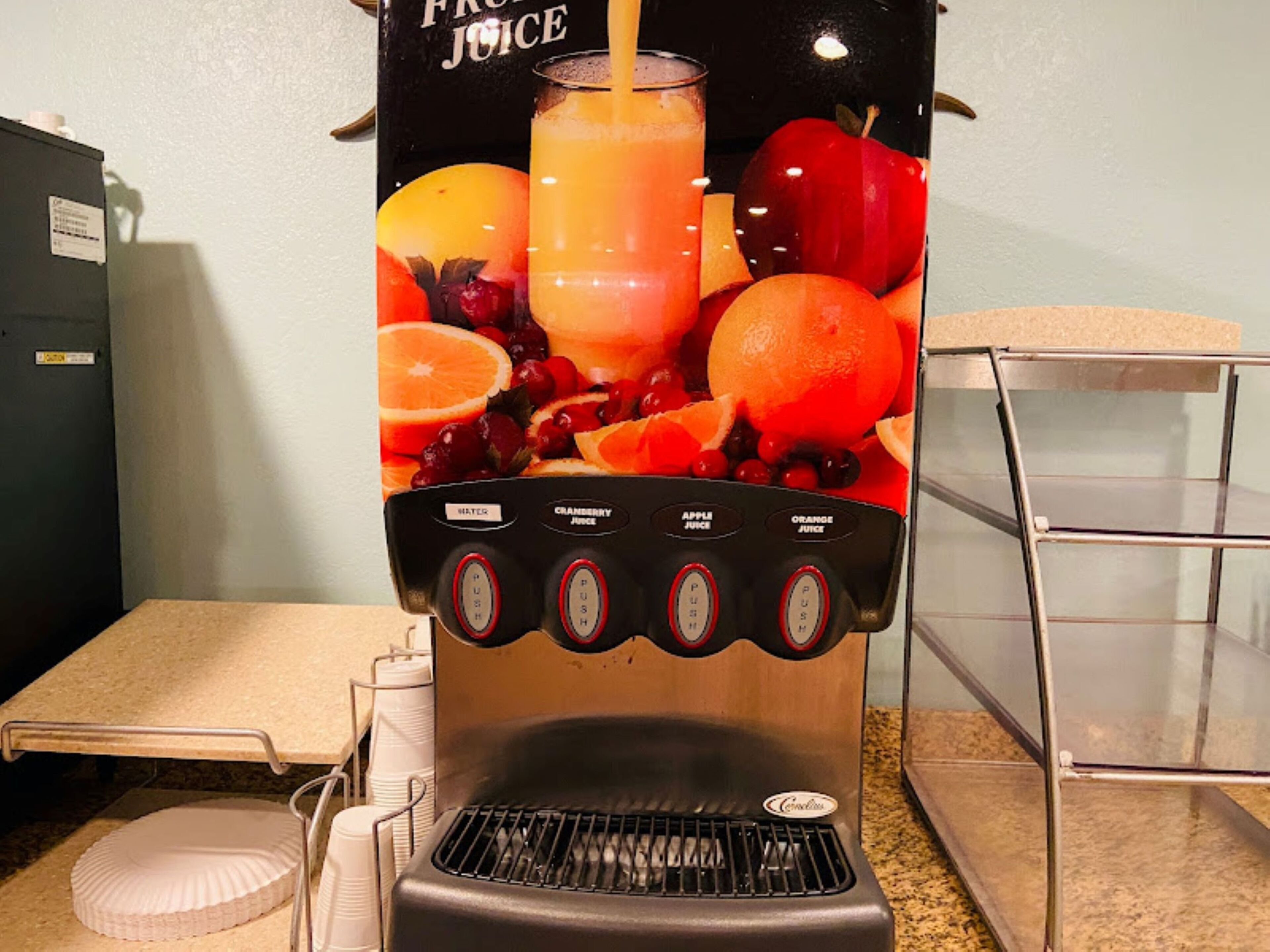 A juice dispenser with multiple push buttons for different types of juice, surrounded by a poster of fruits and a glass of juice.