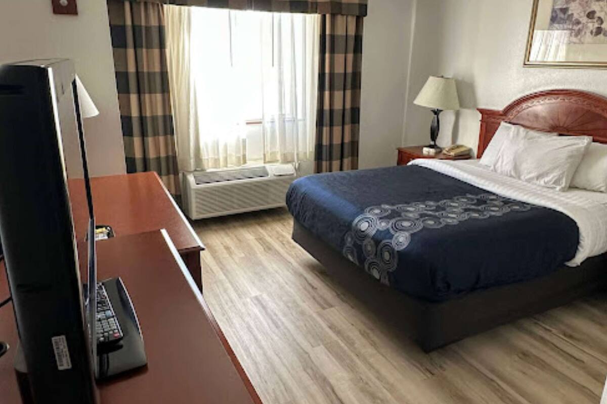 Ett hotellrum med en säng, ett skrivbord med en dator, en tv och ett fönster med gardiner.