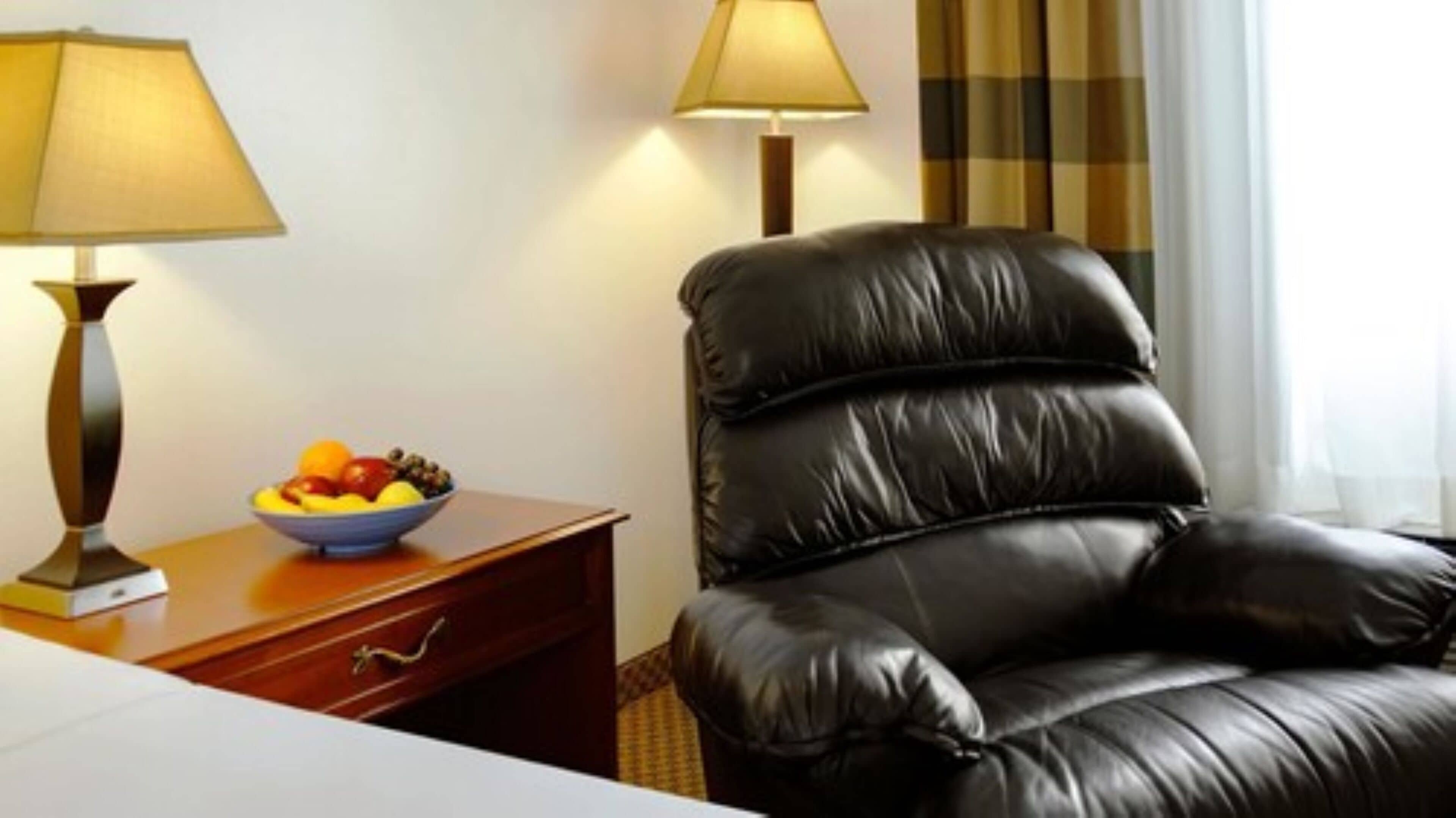 A hotel room with a black leather chair, a wooden nightstand with a bowl of fruit, and two lamps.