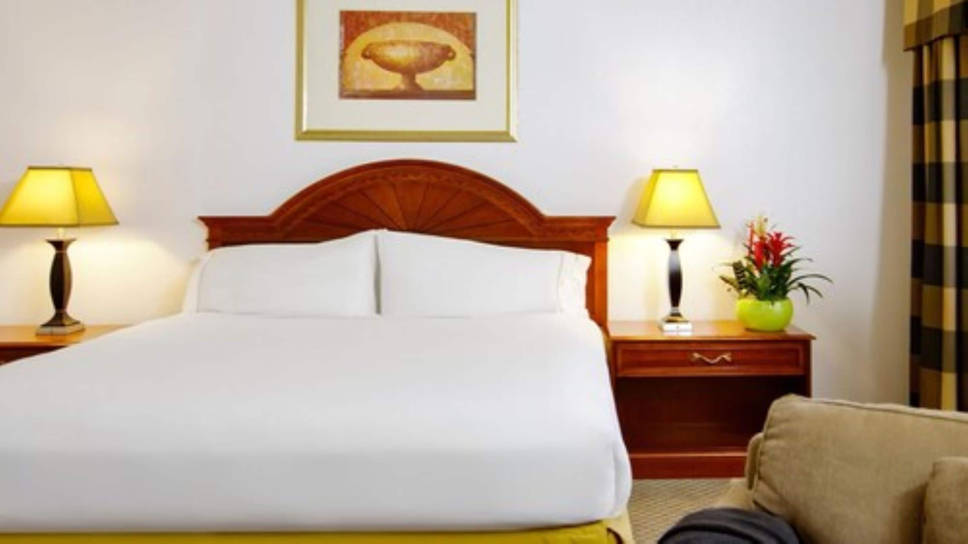 Une chambre d’hôtel avec un grand lit, deux tables de chevet avec des lampes, une petite table de nuit avec un vase et un tableau encadré au mur.