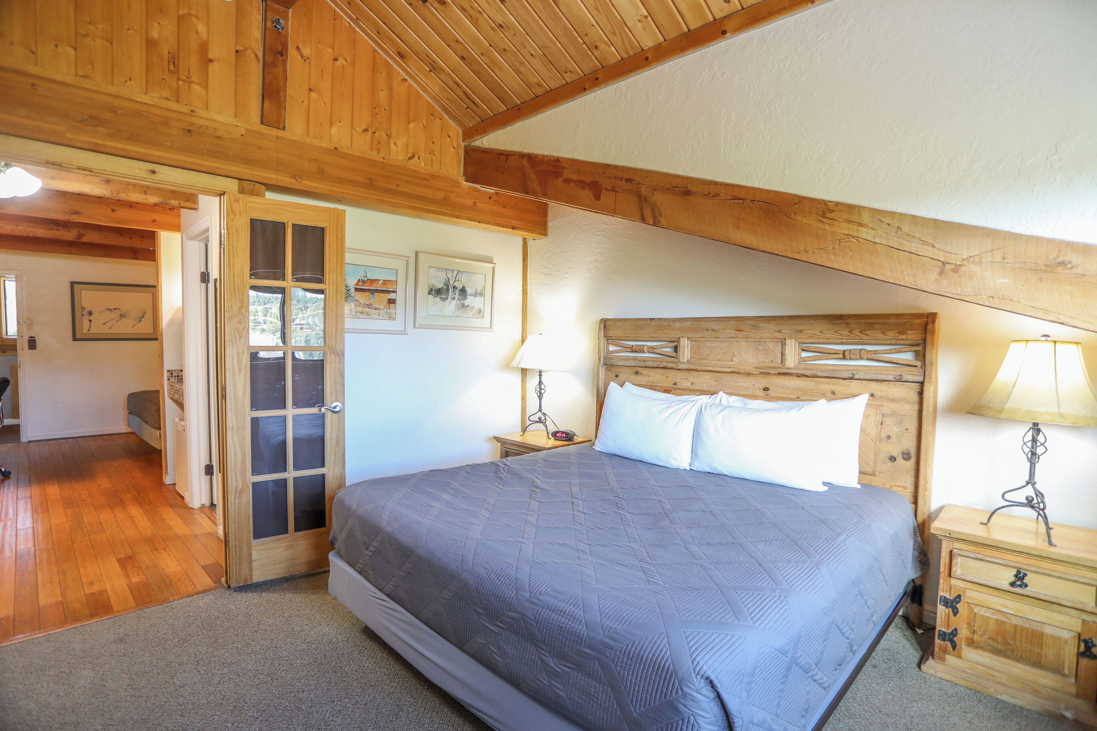 Un dormitorio con techo de madera, una cama grande con colcha gris, dos mesitas de noche con lámparas y una puerta que da a otra habitación.