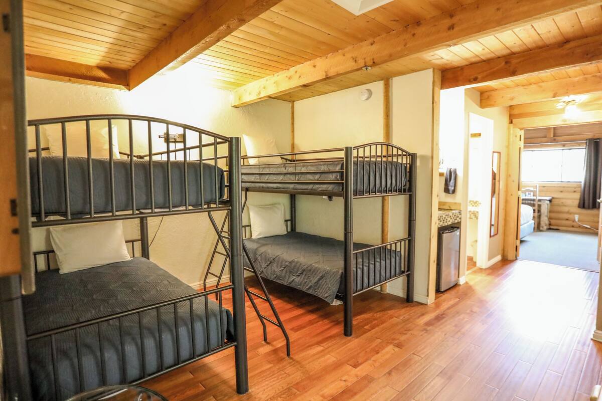A bunk bed room with wooden floors and a kitchen area in the background.