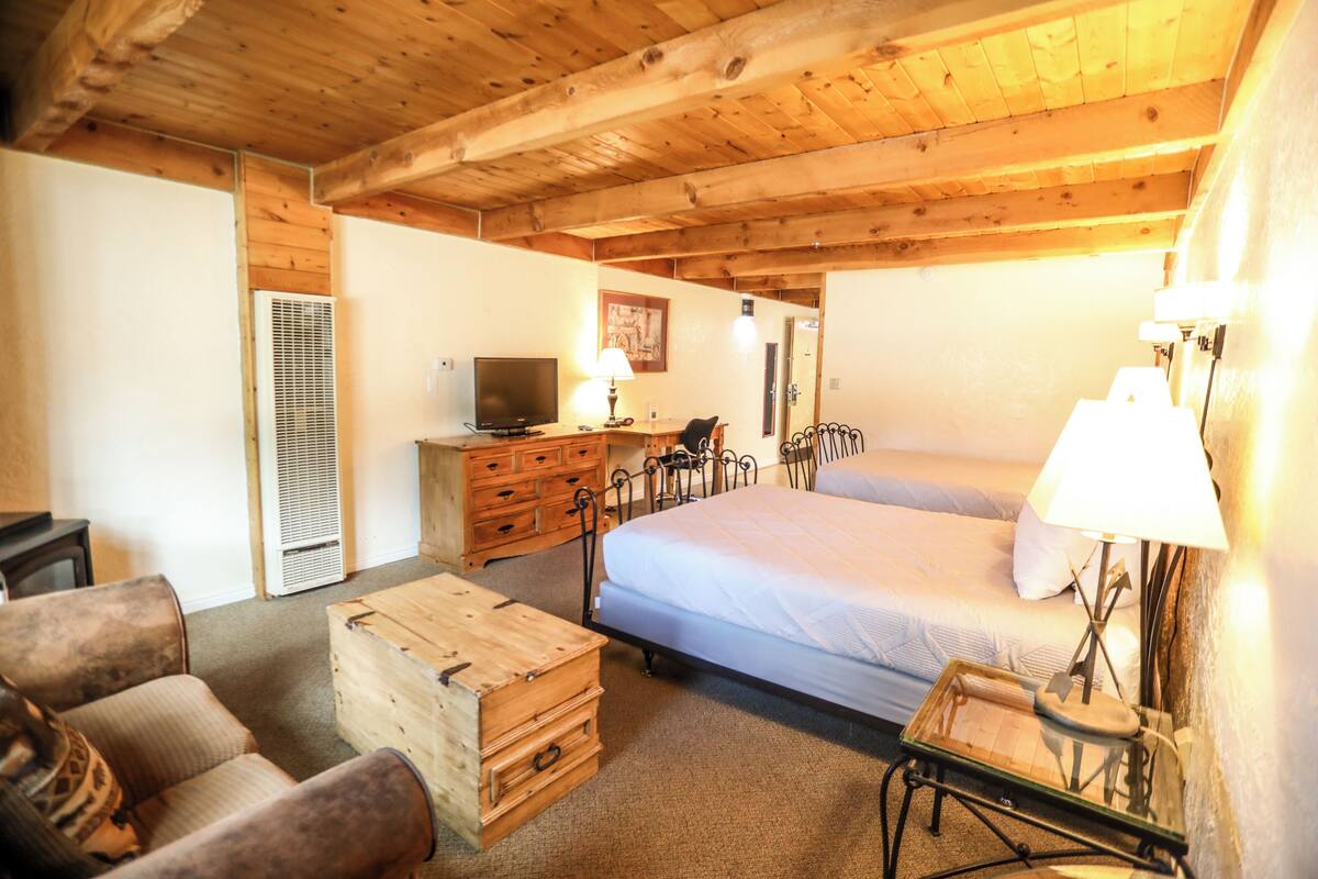 A cozy room with a wooden ceiling, a bed, a desk with a lamp, a television, and a chest of drawers.