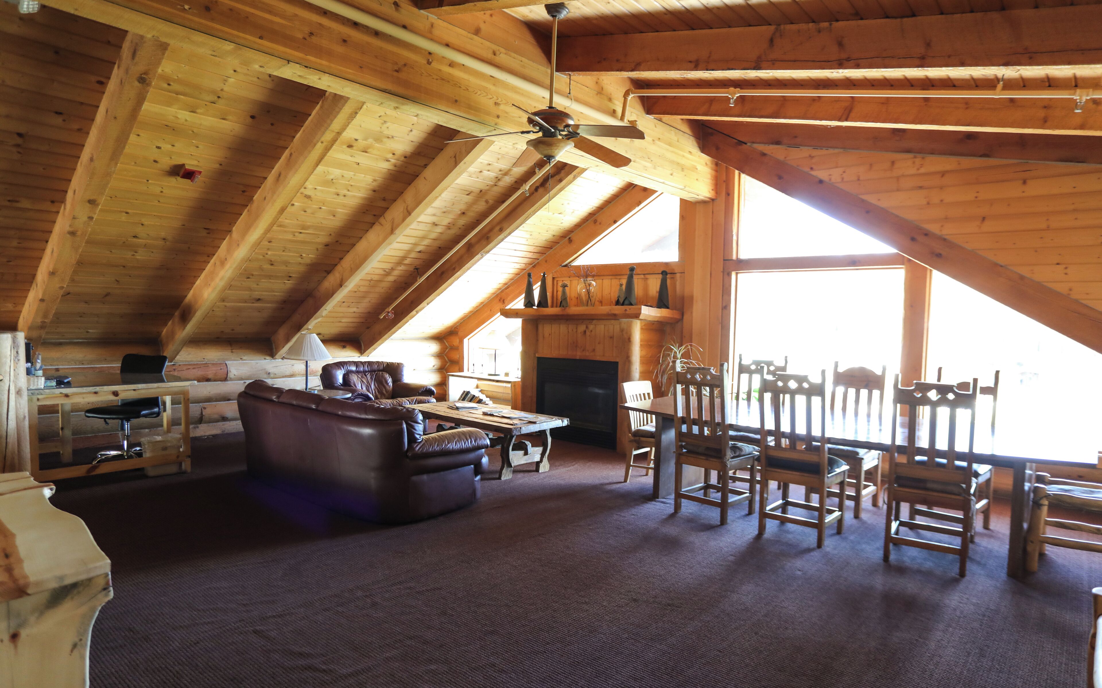 A spacious loft with a high ceiling, wooden beams, a ceiling fan, a fireplace, and a large window.
