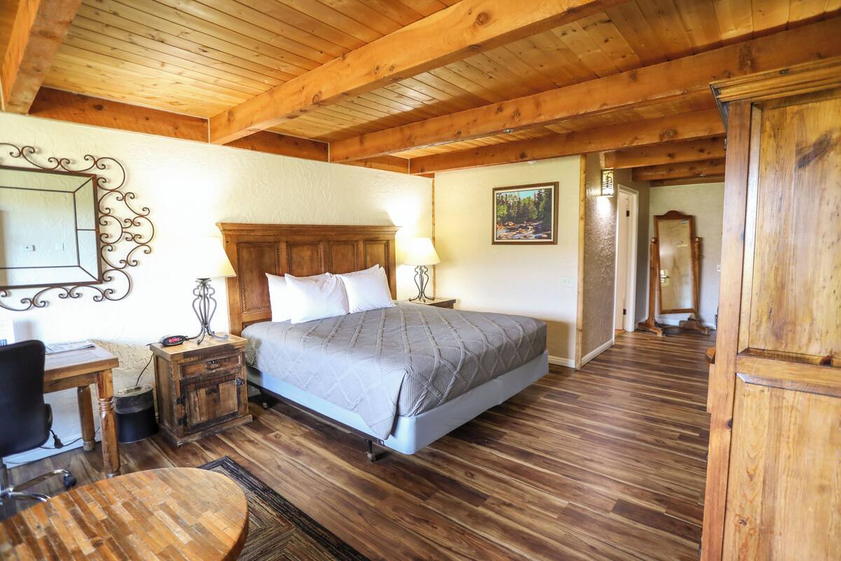 A bedroom with a wooden ceiling, a bed with a grey quilt, a wooden nightstand, a desk with a chair, and a mirror on the wall.