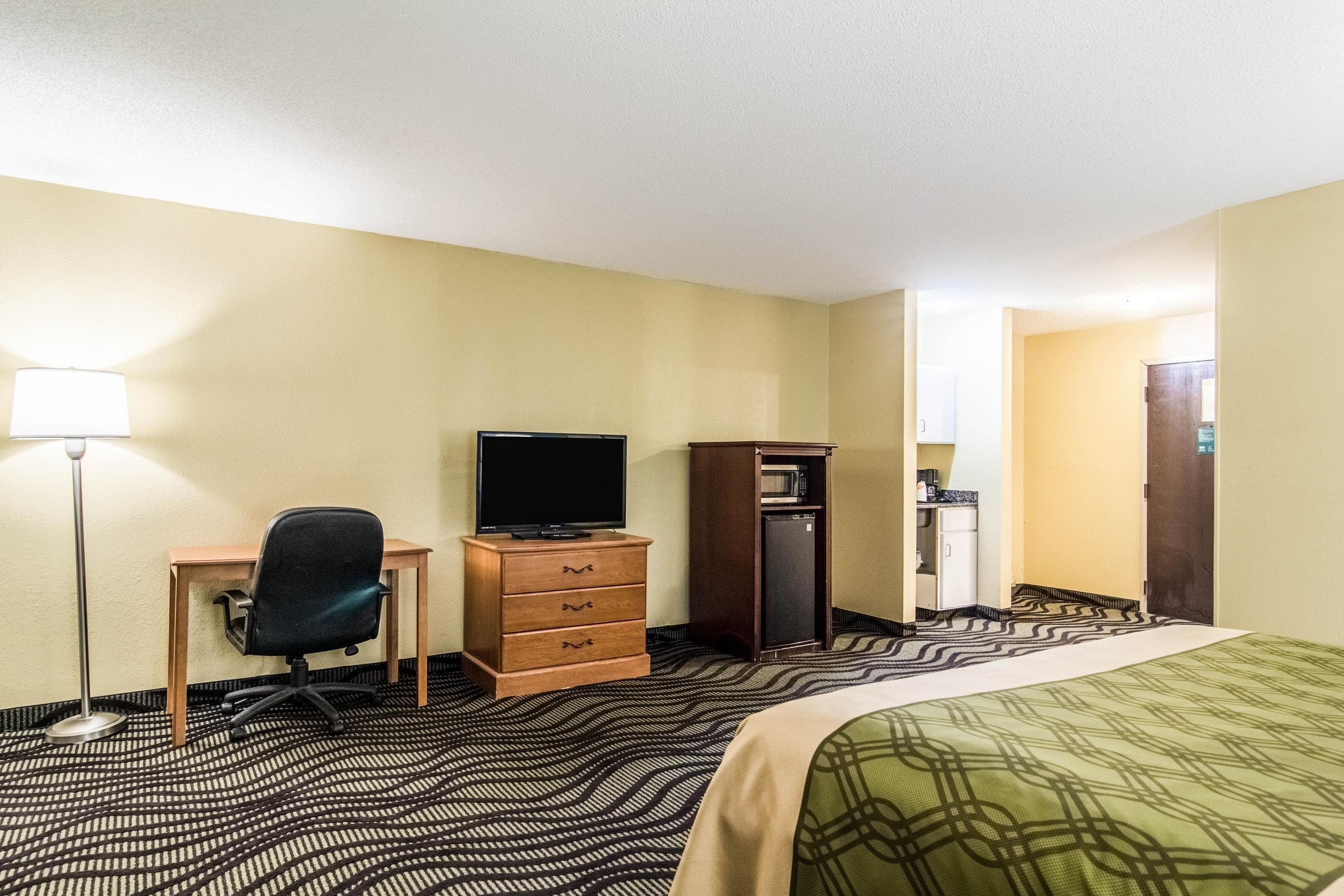 Quarto de hotel com cama, secretária com cadeira, televisão, guarda-roupa e uma kitchenette pequena.
