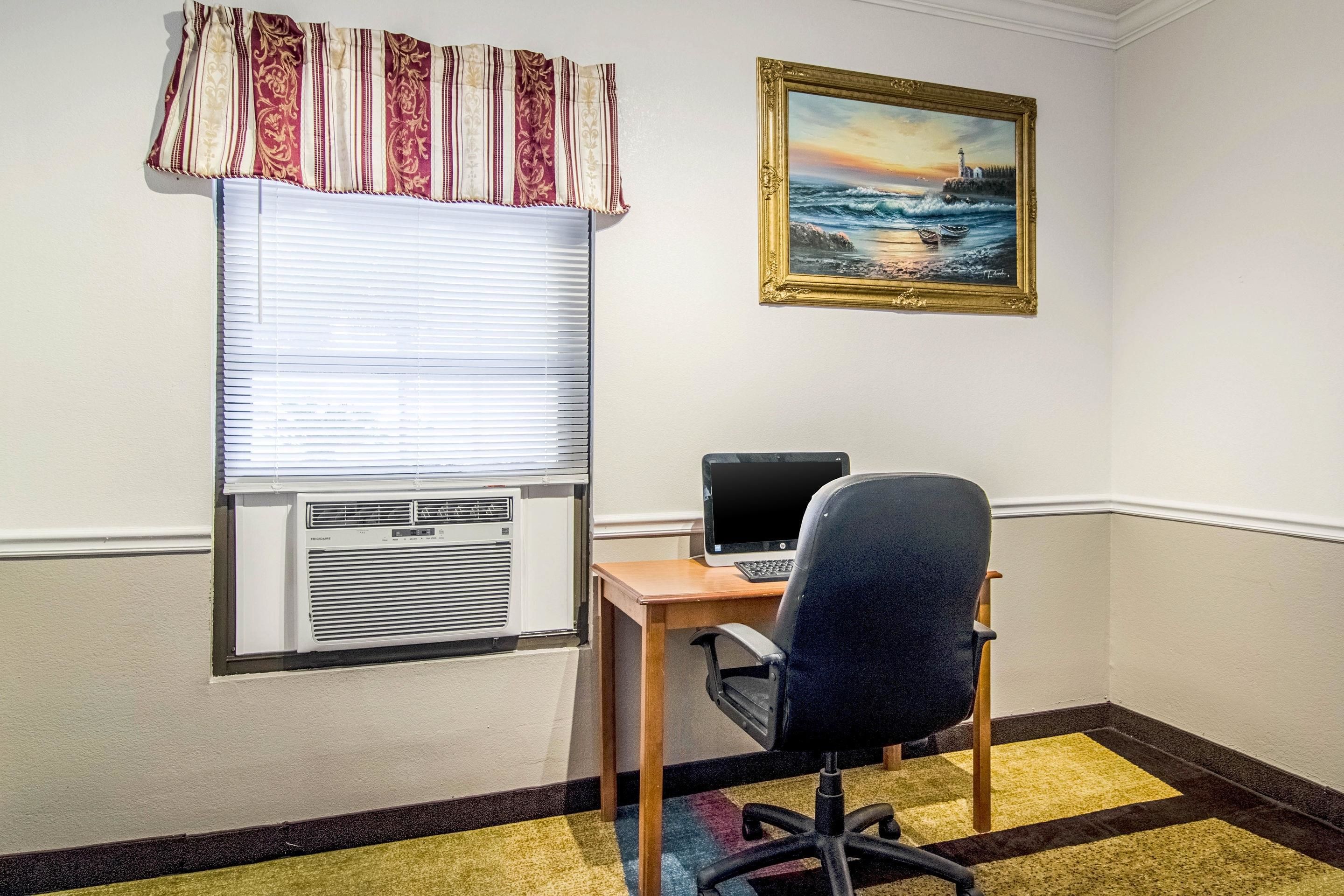 A room with a desk, a chair, a window with blinds, an air conditioner, and a painting of a lighthouse on the wall.