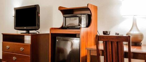 A compact hotel room with a wooden desk, a television, a microwave, a refrigerator, a chair, and a lamp.