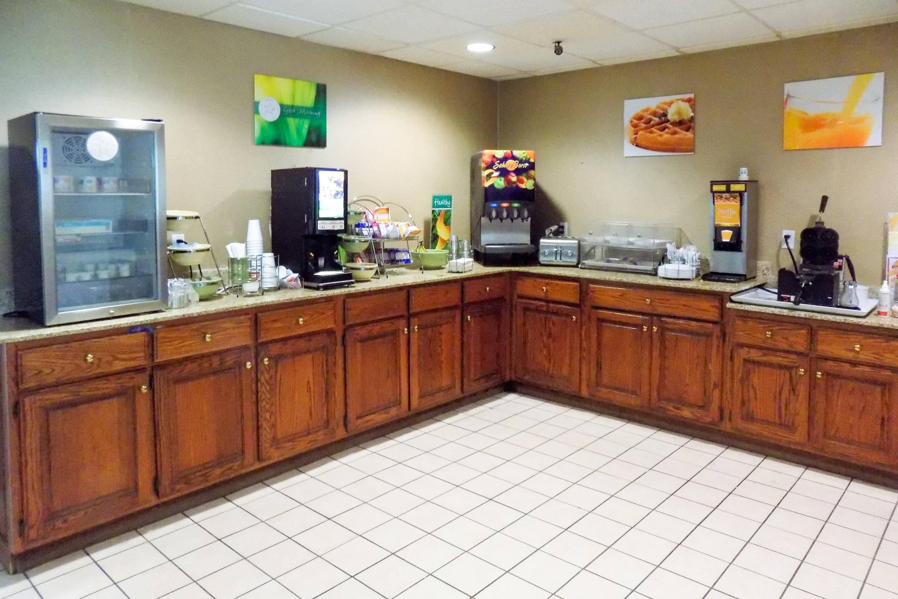 A hotel kitchen with wooden cabinets, a refrigerator, a coffee machine, and a waffle maker.