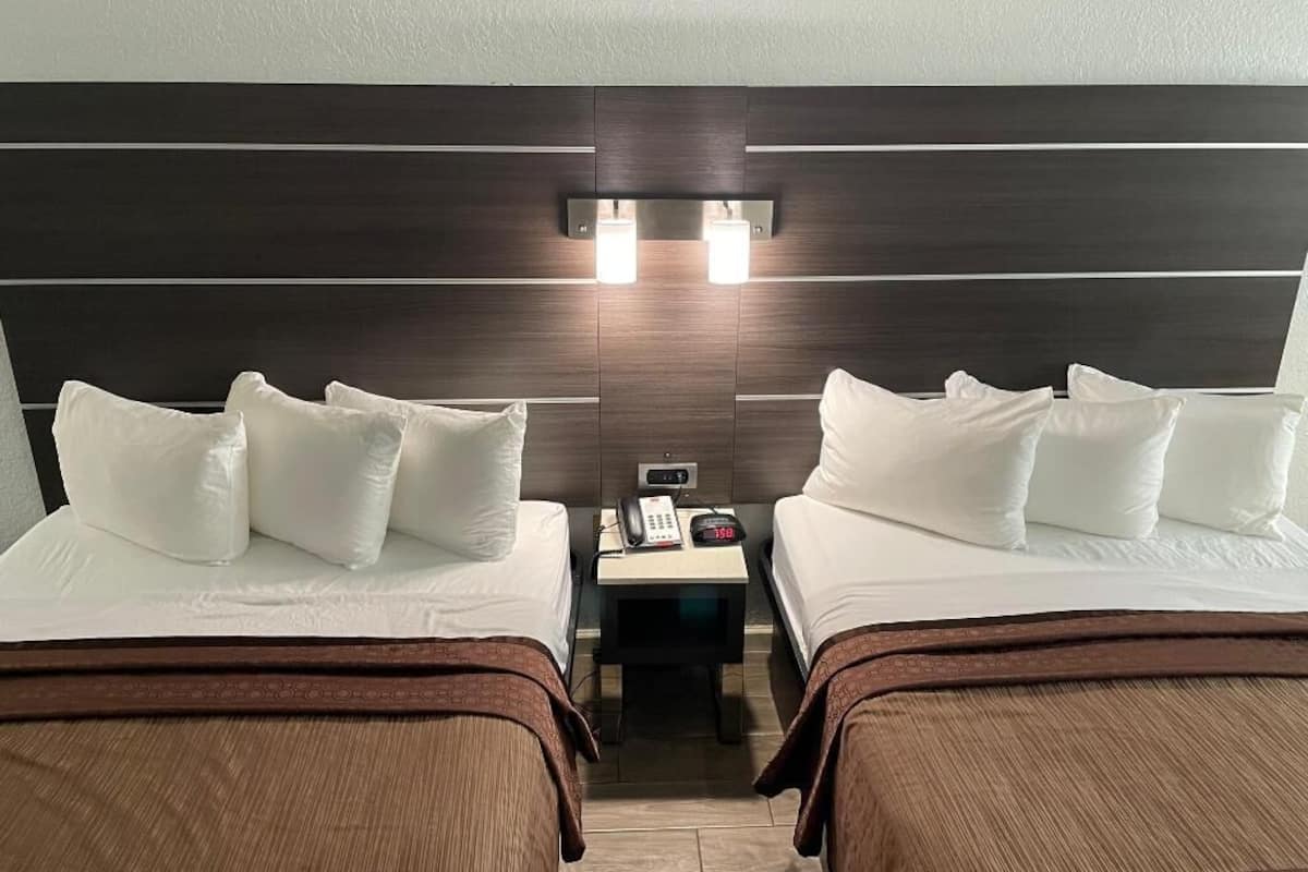 A hotel room with two beds, a nightstand with a phone and a clock, and a wall-mounted light fixture.
