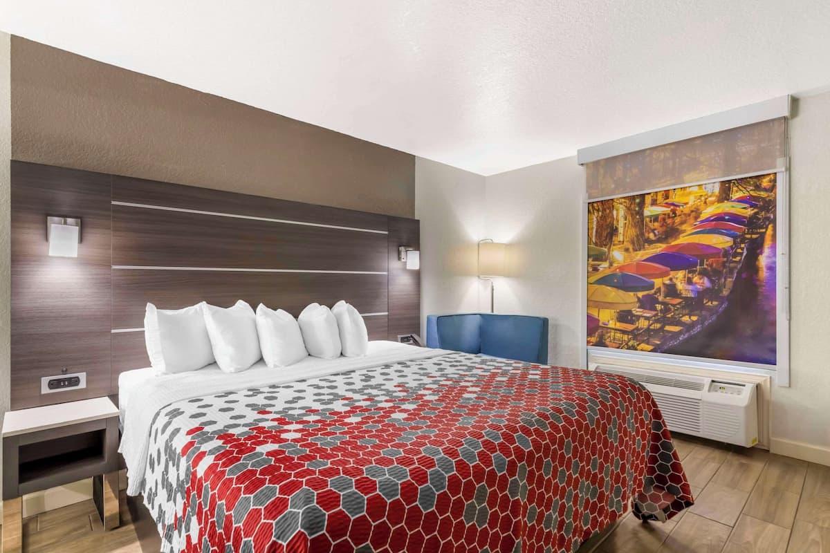 A hotel room with a large bed, a nightstand, a desk, a blue chair, a wall-mounted lamp, and a large window with a colorful view.