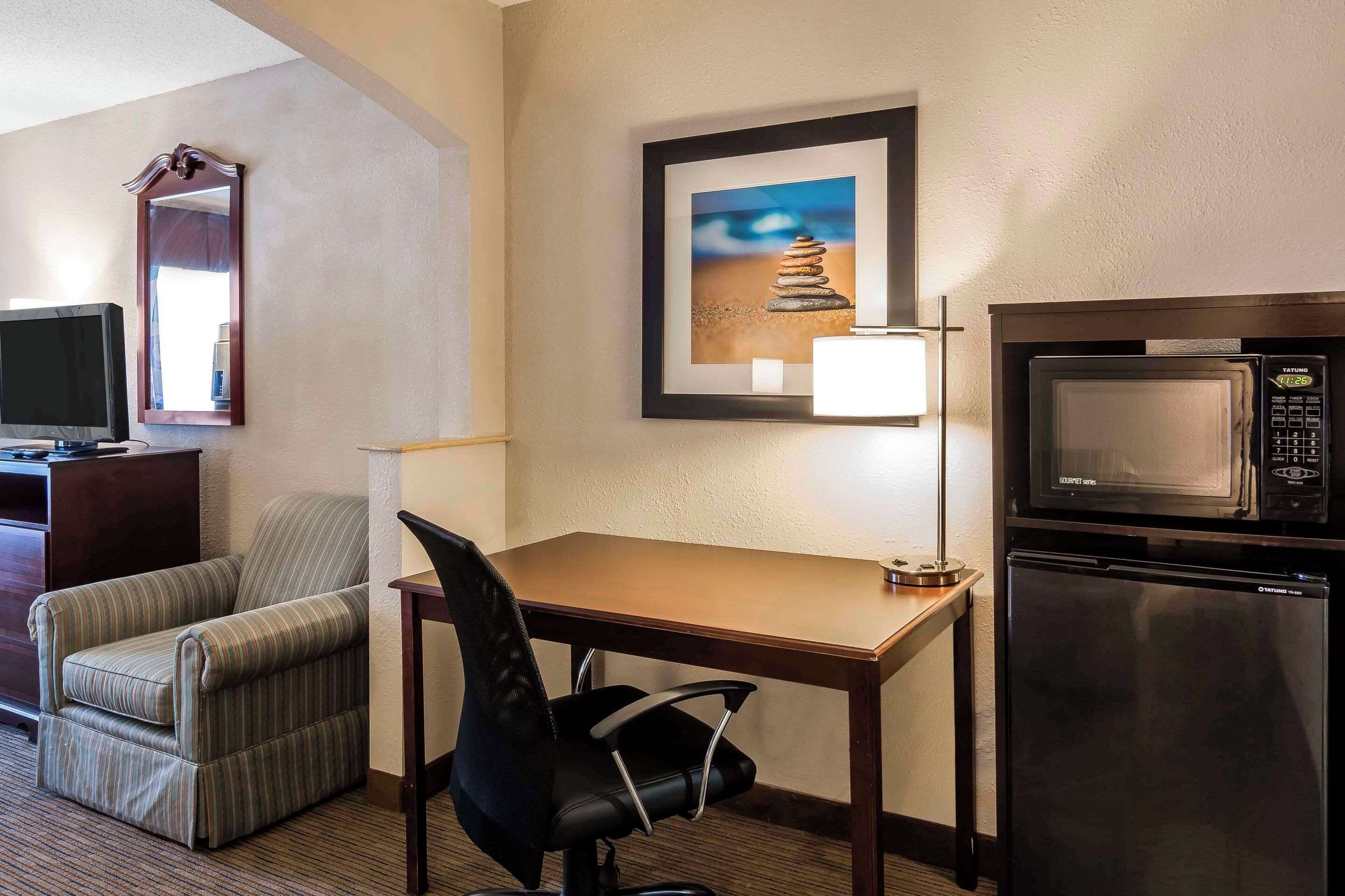 A hotel room with a wooden desk, a black office chair, a microwave, a refrigerator, a television, and a framed picture of a stack of stones on the wall.