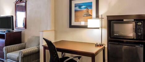 A hotel room with a wooden desk, a black office chair, a microwave, a refrigerator, a television, and a framed picture of a stack of stones on the wall.