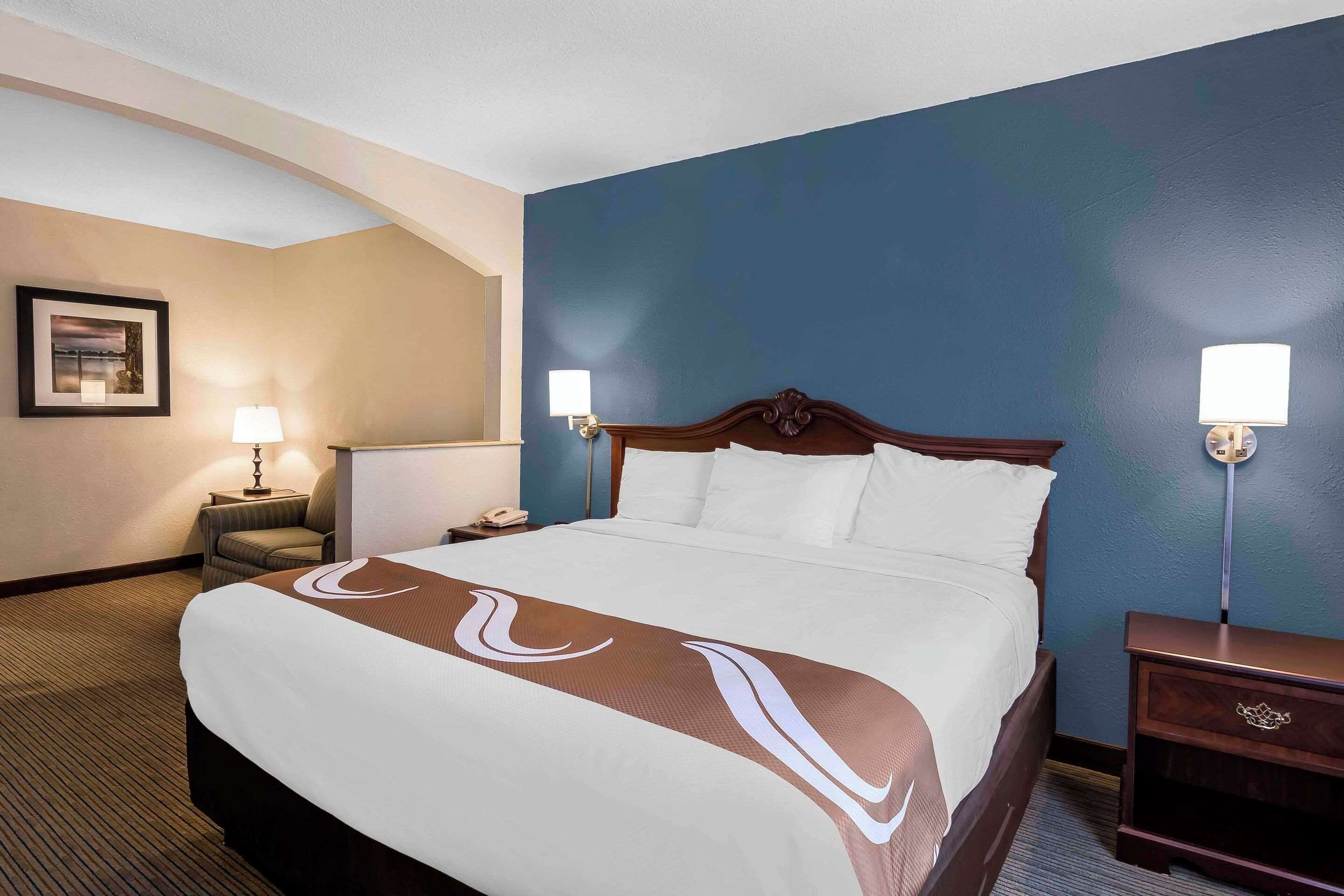 Habitación de hotel con una cama grande, dos lámparas de noche, una mesita de noche, una silla y un cuadro en la pared.
