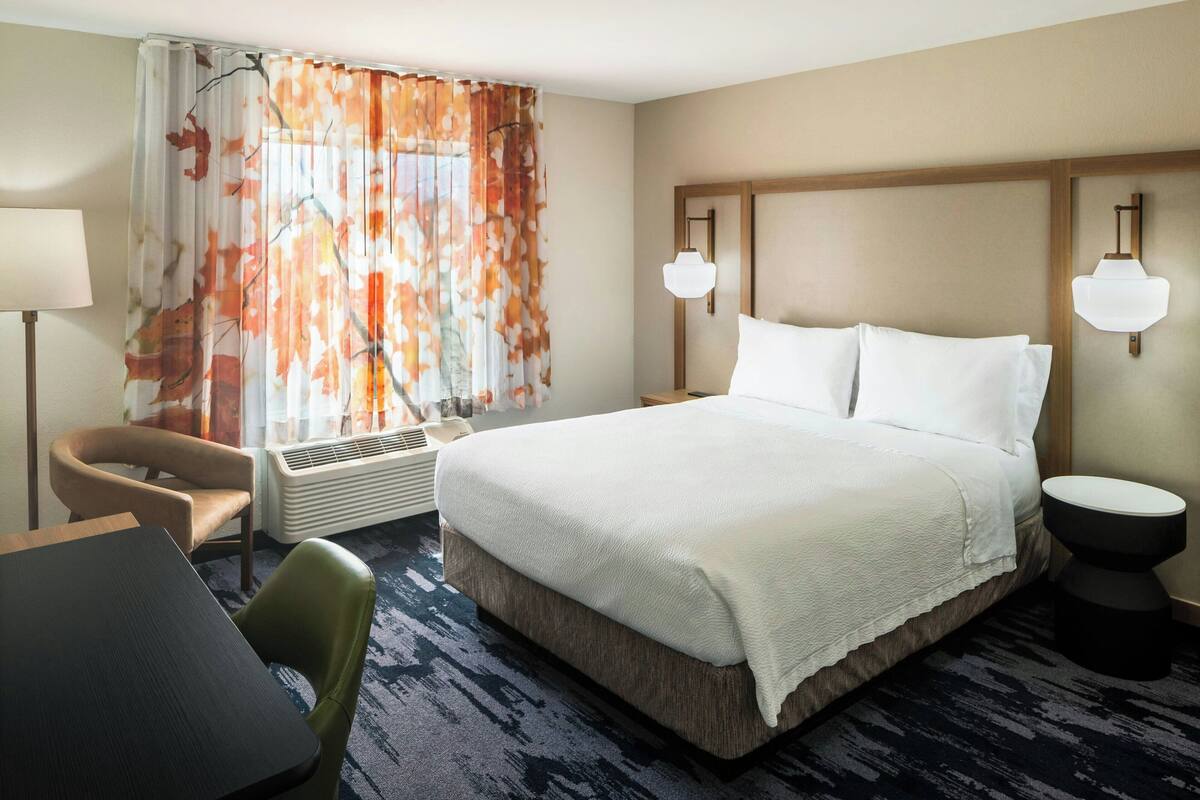 A hotel room with a bed, a chair, a table, a lamp, and a heater.
