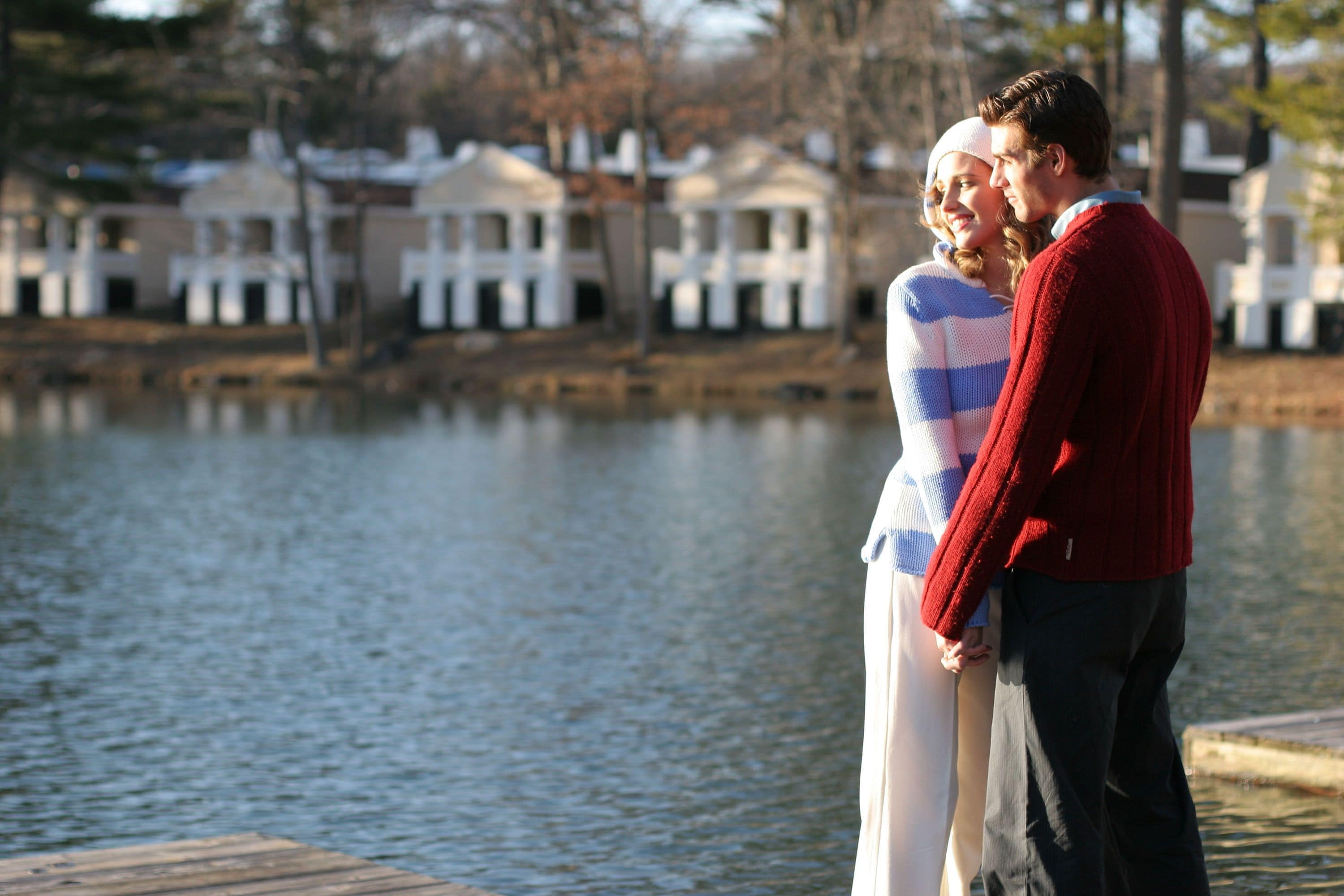 A couple standing by a lake with a row of houses in the background.