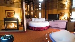 A hotel room with a fireplace, a large bed with a red skirt, a TV, and a bathtub with candles. - Paradise Stream Resort (Cresco)