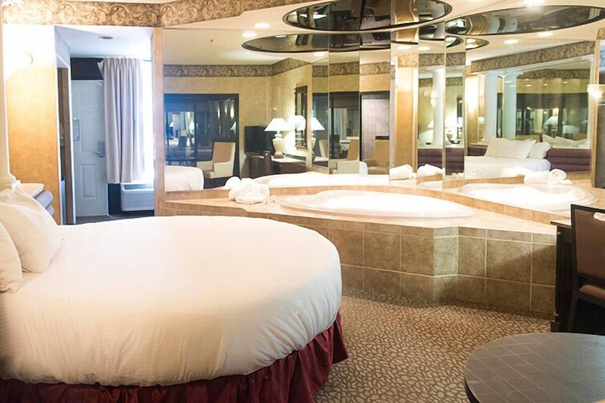 A spacious hotel room with a large bed, a separate sitting area, a bathtub, and a large mirror reflecting the room.