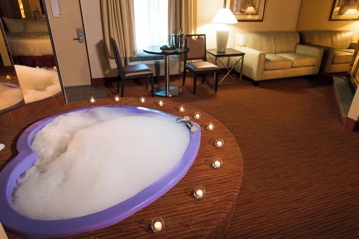 A hotel room with a large bathtub, a dining area, and a living area with a sofa and lamp.