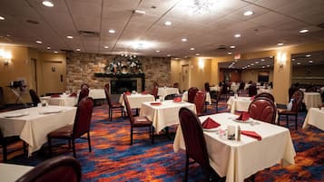 A banquet hall with multiple tables and chairs, a stone wall with a fireplace, and a carpeted floor.