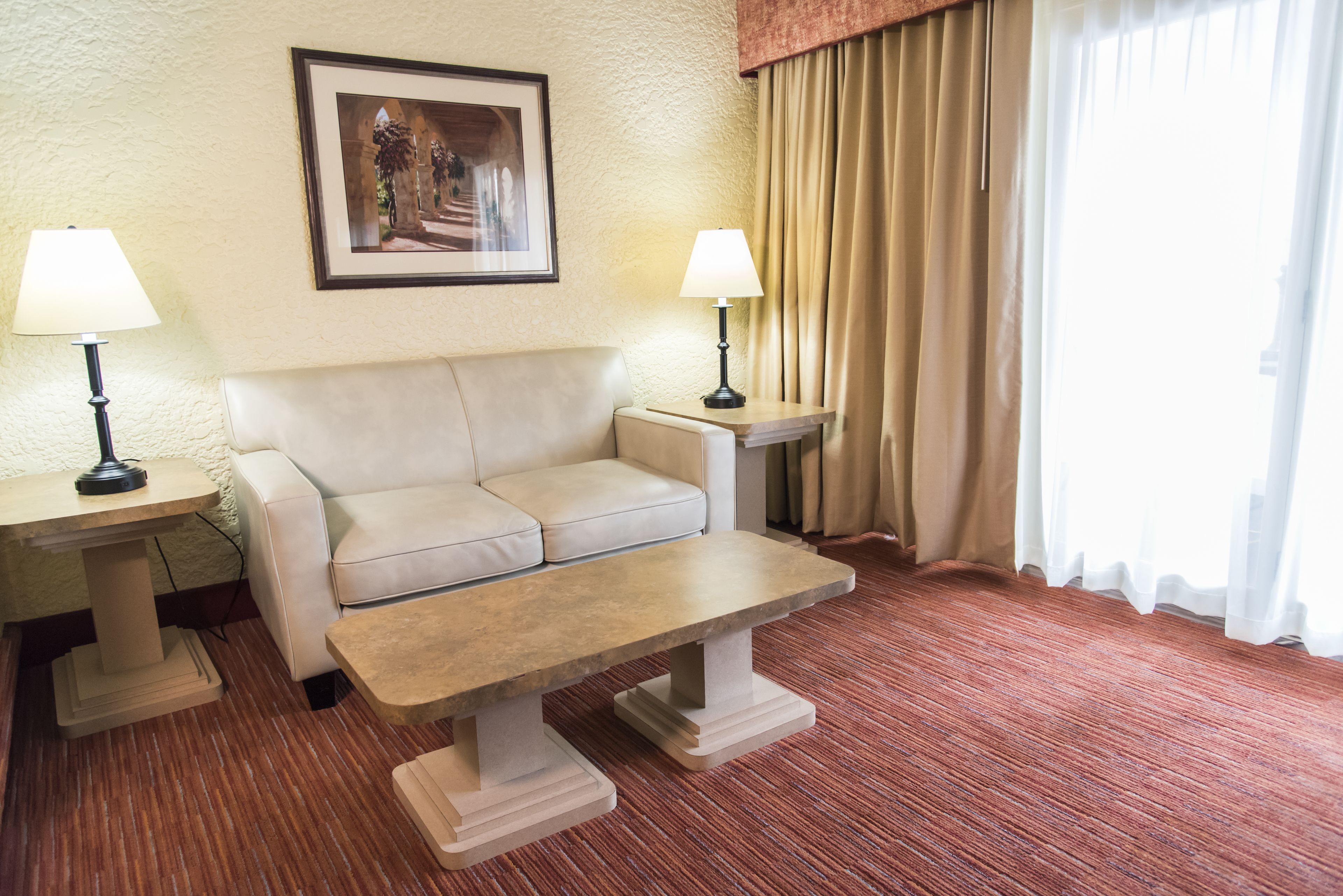 A hotel room with a beige sofa, two bedside tables with lamps, a framed picture on the wall, and a window with curtains.