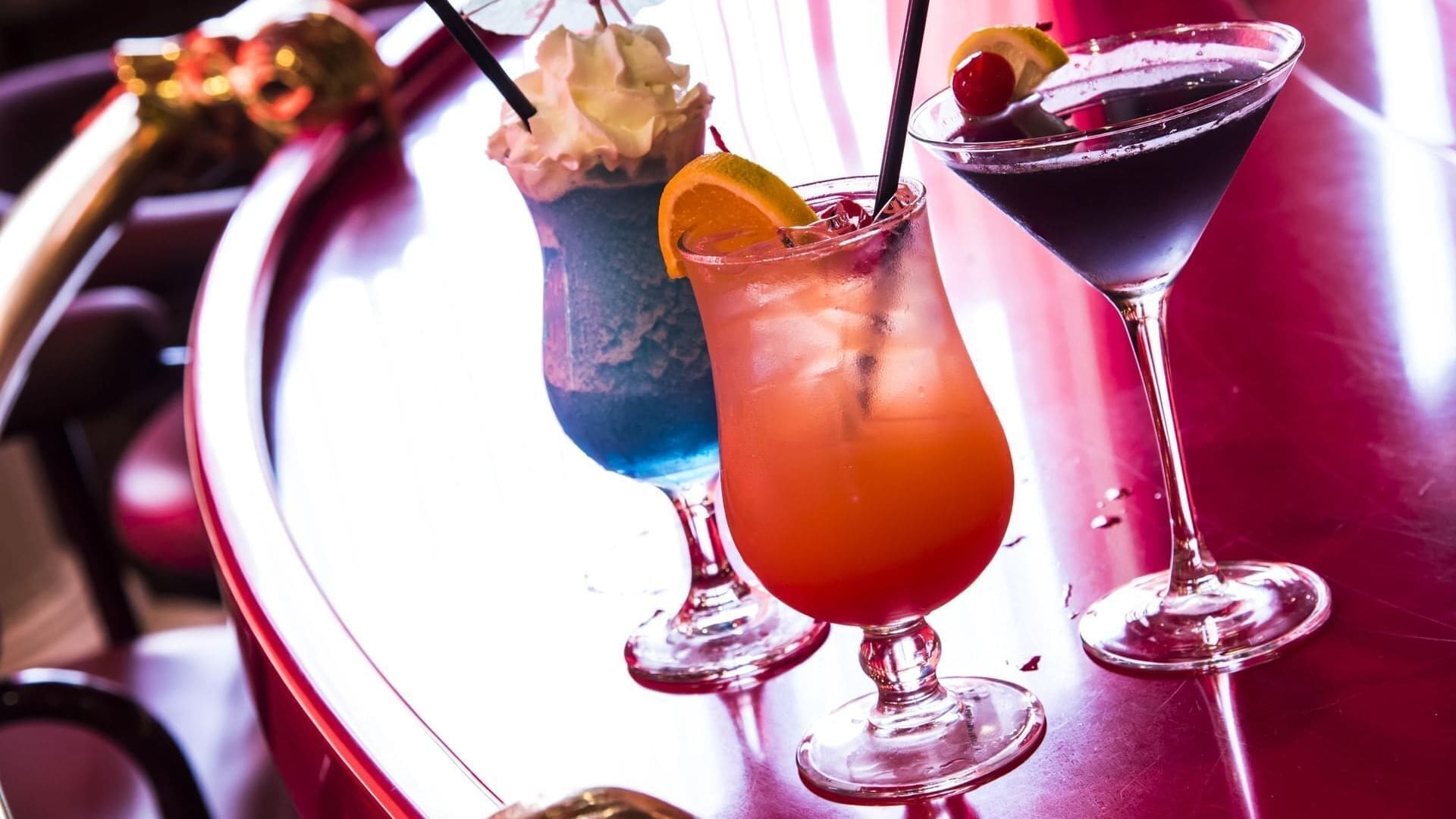 A bar area with three drinks in martini glasses, one with a blue drink and two with red drinks, each garnished with a slice of orange and a cherry.