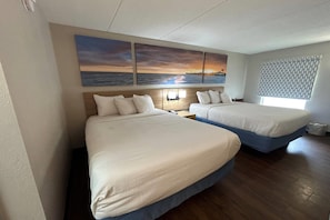 A hotel room with two beds, a large wall painting of a sunset over the ocean, a window with a patterned blind, and a wooden floor. - Days Inn by Wyndham Ridgeland South Carolina (Ridgeland)