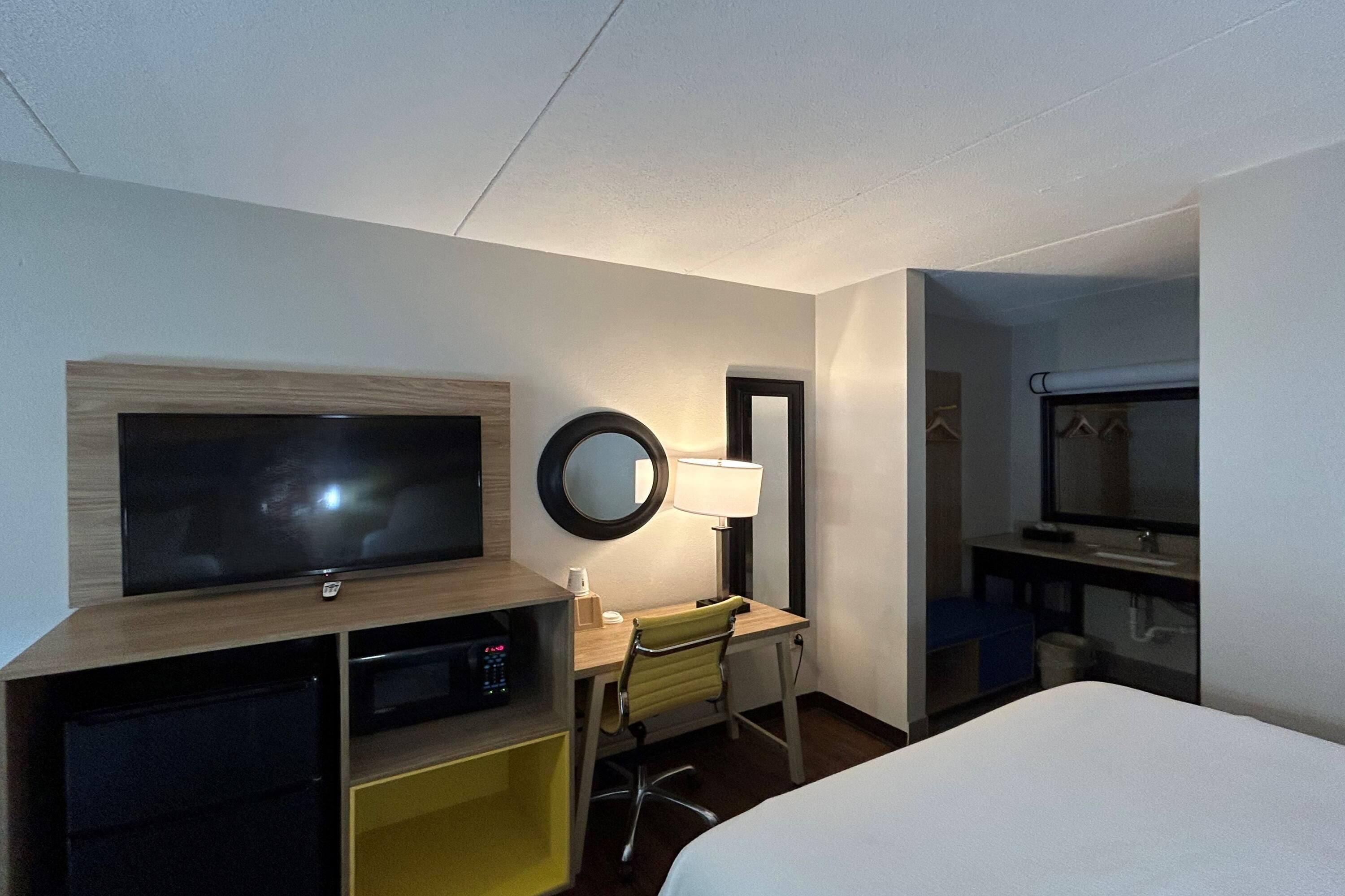 A hotel room with a bed, a desk with a chair, a television, a mirror, and a window.