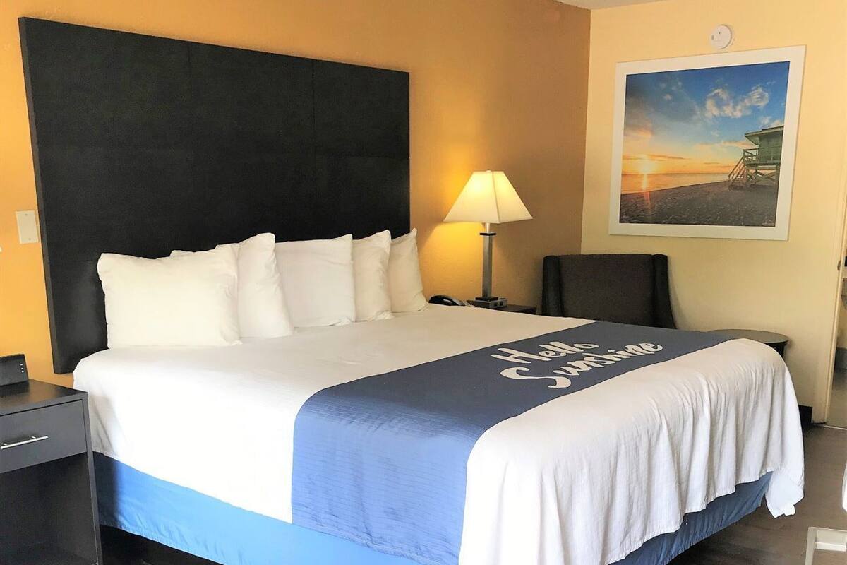 A hotel room with a bed, a nightstand, a lamp, a wall clock, and a framed picture.