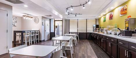 Een modern restaurantinterieur met witte tafels en stoelen, een lange toonbank met een koffiemachine, en een geel accentmuur.