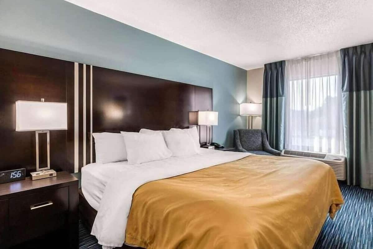 A hotel room with a bed, bedside lamps, a chair, a desk with a clock, and a window with curtains.