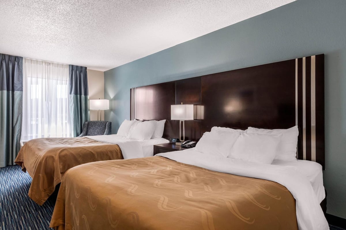 A hotel room with two beds, a large dark headboard, a desk with a lamp, and a chair.