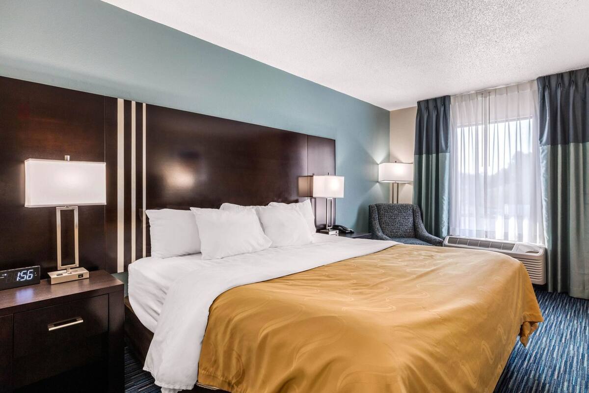 A hotel room with a bed, bedside lamps, a chair, a desk with a phone, and a digital clock.