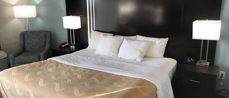 Et hotelværelse med en stor seng, to sengeborde, en lænestol og et natbord med telefon og digitalt ur.