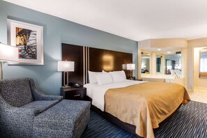 A hotel room with a large bed, a sofa, a desk with a lamp, a mirror, and a framed picture on the wall.