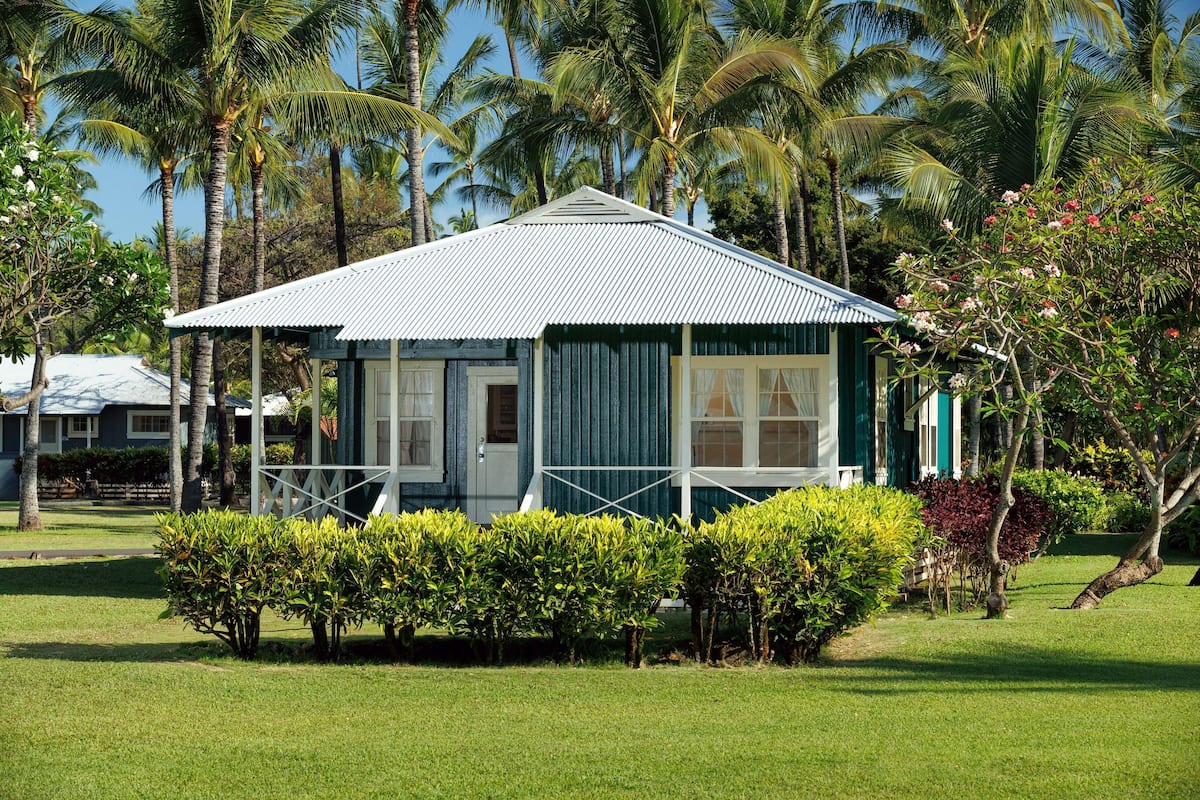 A small, single-story building with a corrugated metal roof, surrounded by well-maintained grass and various tropical plants.