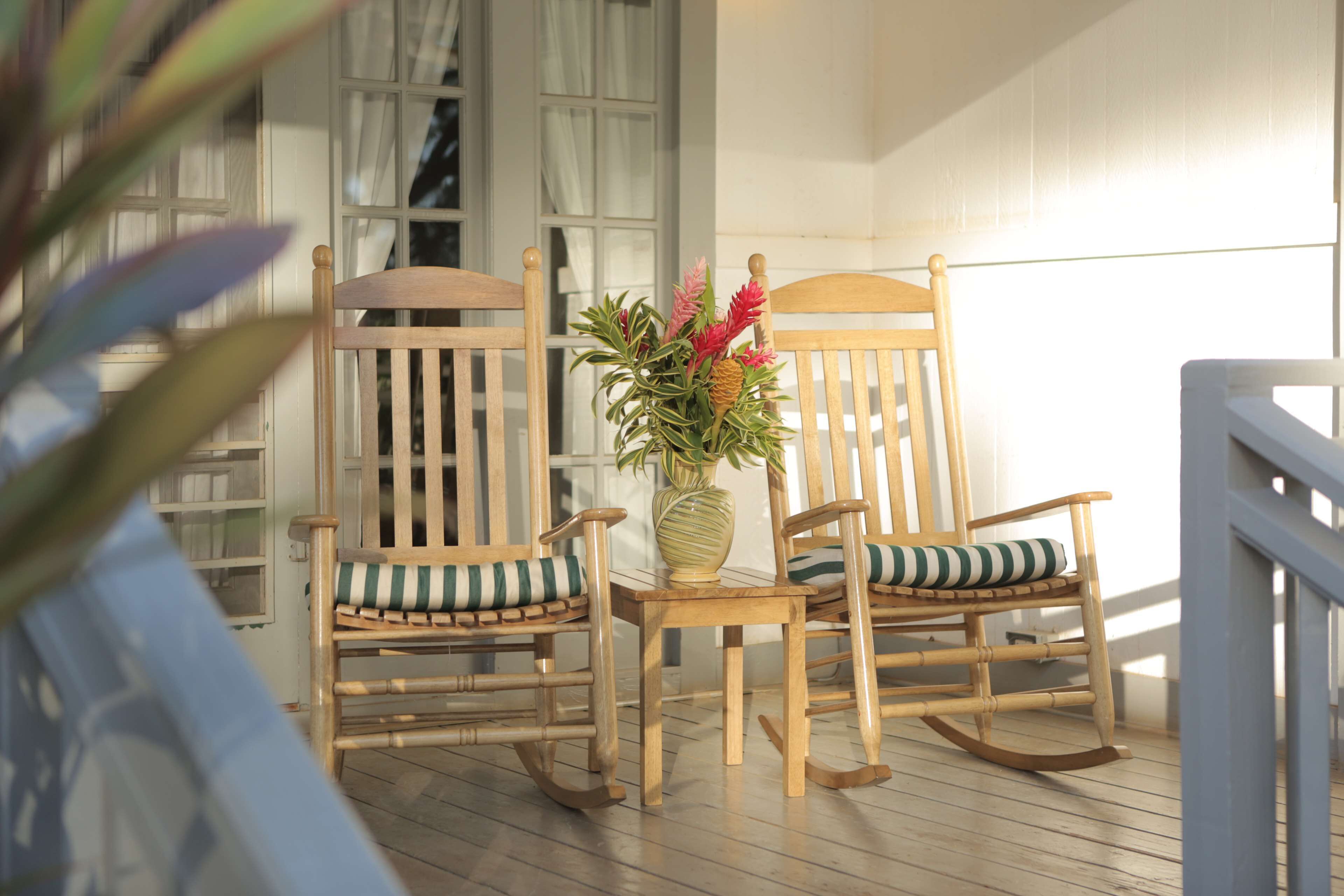 Two rocking chairs with striped cushions on a wooden deck, a vase with flowers, and a window with white curtains.