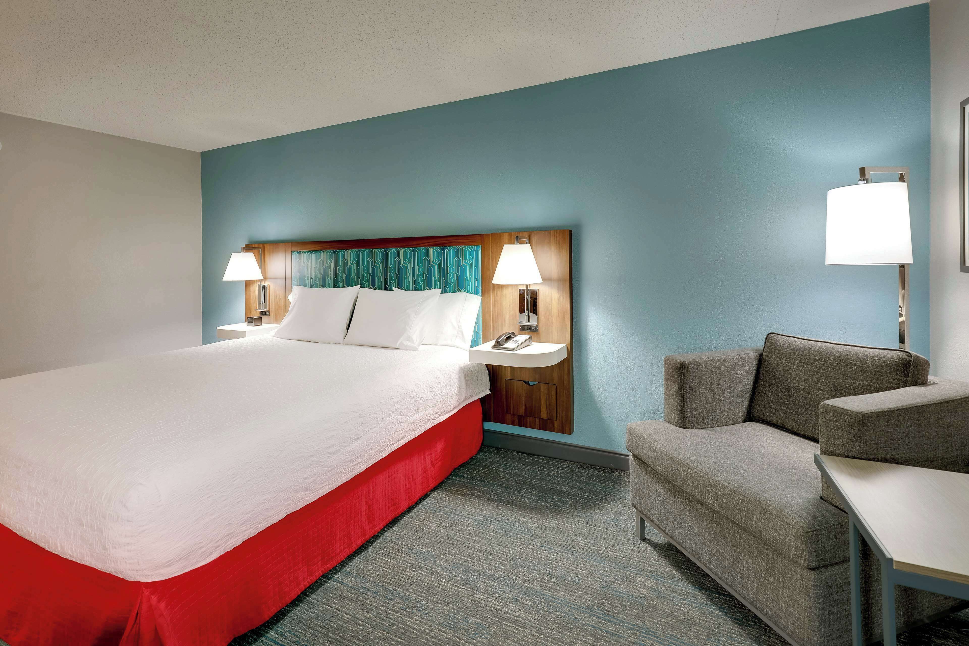 A hotel room with a large bed, two bedside tables with lamps, a grey armchair, and a small table.