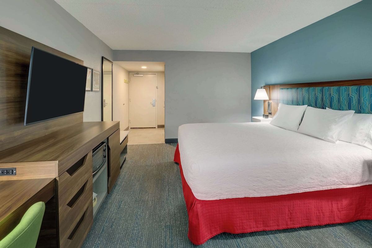 A hotel room with a bed, a desk with a television, a chair, and a door leading to another room.