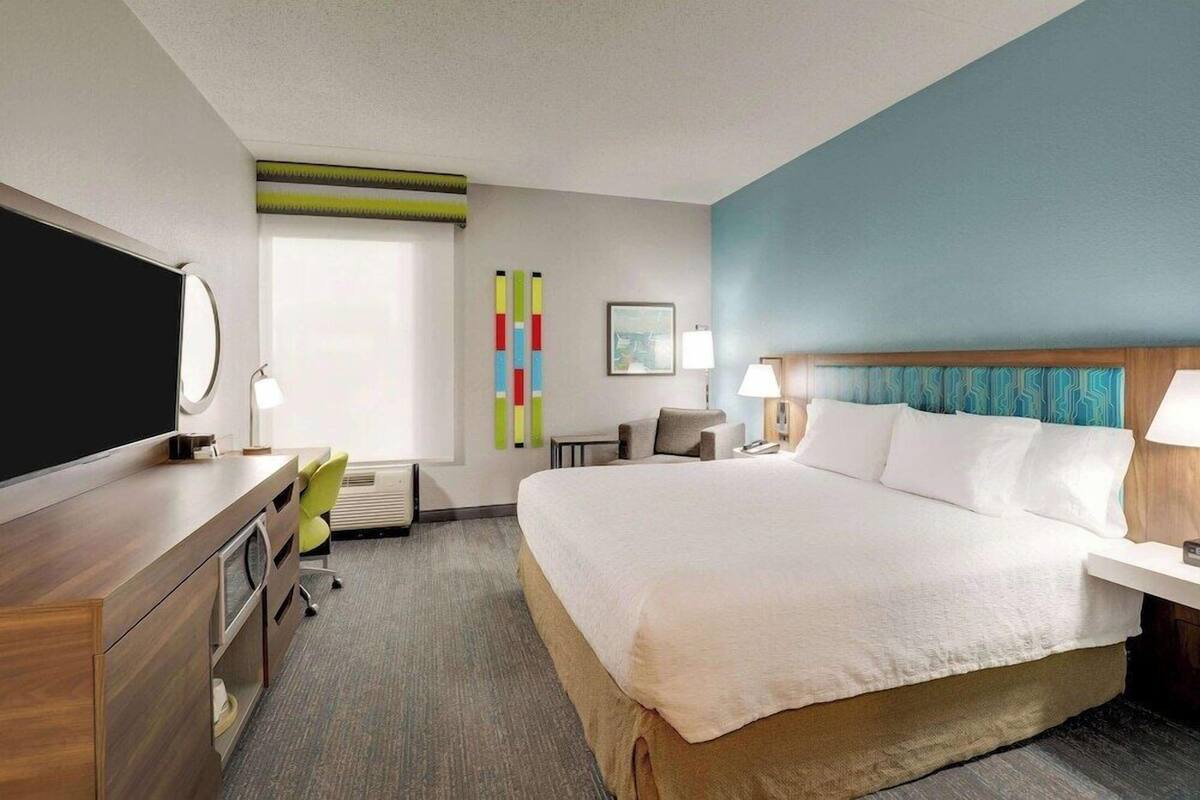 A hotel room with a large bed, a desk with a flat-screen TV, a chair, a desk lamp, a wall-mounted TV, a window with blinds, and a colorful abstract art piece.
