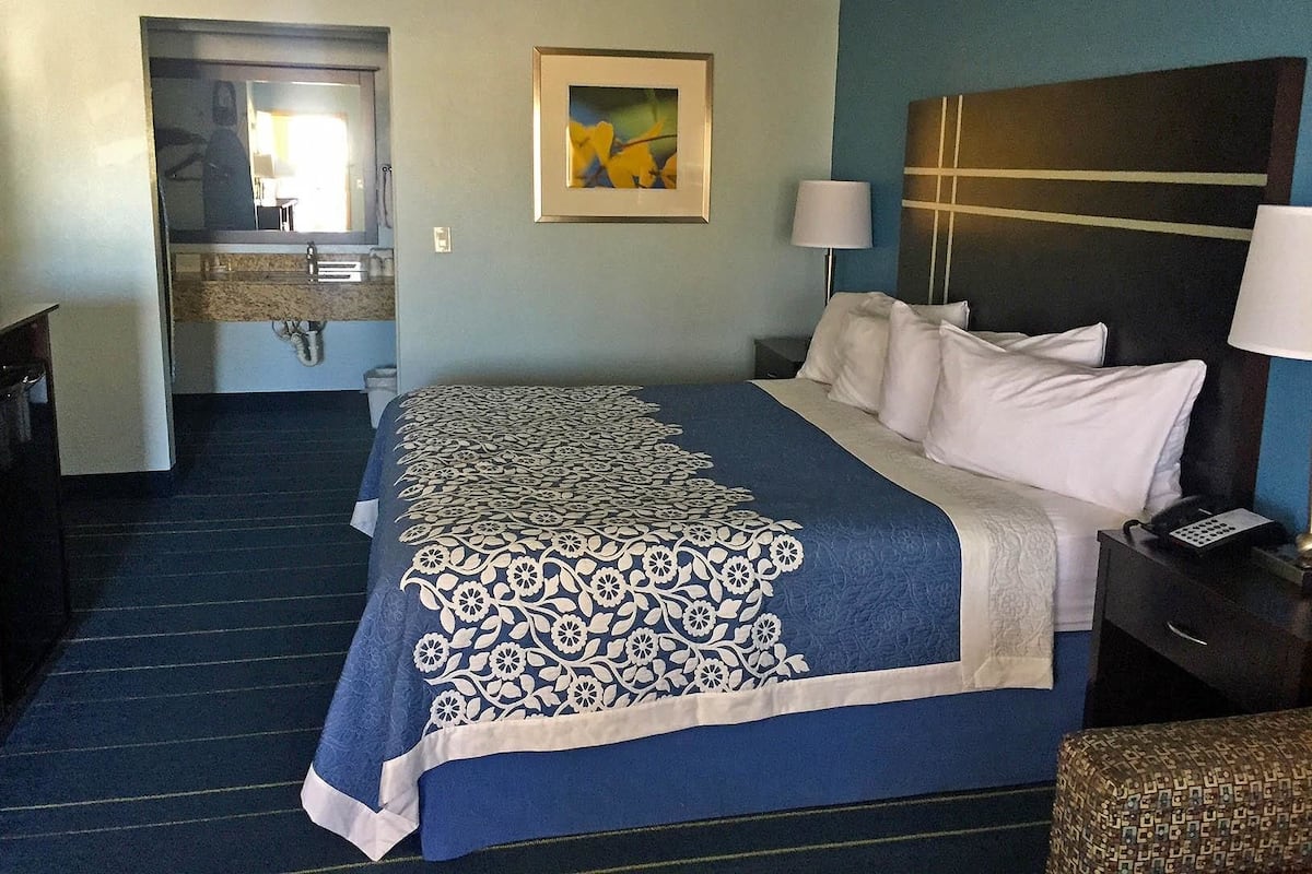 A hotel room with a bed, a nightstand, a lamp, a painting on the wall, and a bathroom visible through an open door.