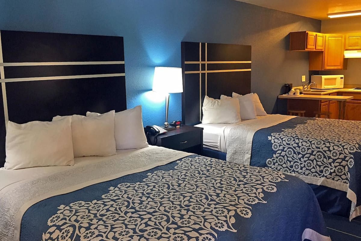 A hotel room with two beds, a nightstand, a lamp, a microwave, and a small kitchen area.