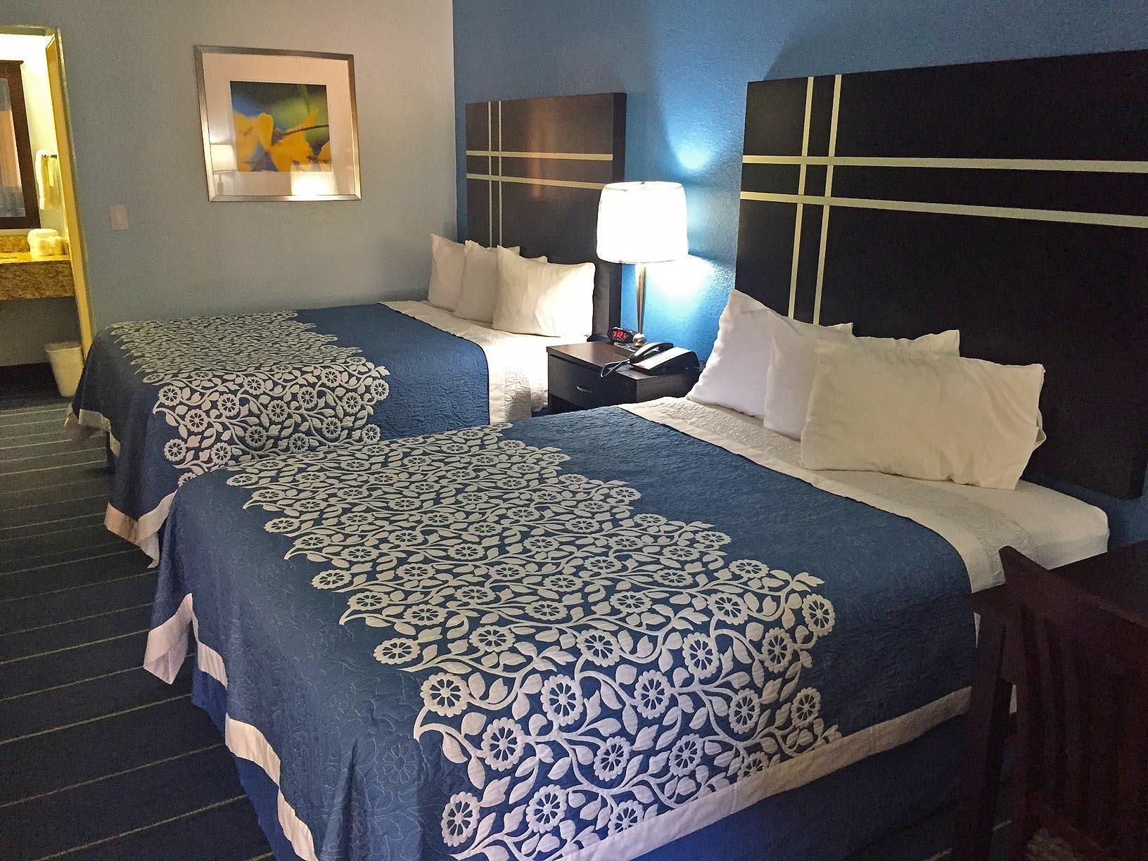 A hotel room with two beds, each with a blue and white patterned bedspread, a bedside table with a lamp, and a framed picture on the wall.